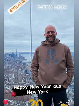 Ein bärtiger Mann in einem braunen Kapuzenpullover und Brille.