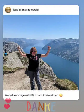 Eine Frau in einem schwarzen T-Shirt und grauen Hosen steht auf einer Klippe mit erhobenen Armen und blickt auf einen Fjord.