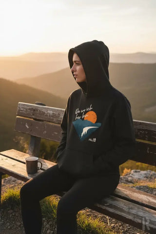 Schwarzer Hoodie mit einem Grafikdruck einer orangenen Sonne über blauen Bergen und weißen Wellen, mit dem Text ’du wirst hier bleiben’ darüber.