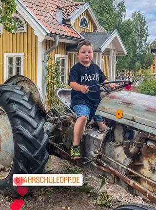 Ein junger Junge in einem schwarzen T-Shirt und blauen Shorts sitzt auf einem alten, grauen Traktor.