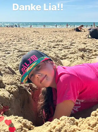 Ein junges Mädchen mit langen braunen Haaren, das eine schwarze und multi-colorierte Baseballmütze und ein leuchtend pinkes T-Shirt trägt, bis zur Brust im Sand begraben.