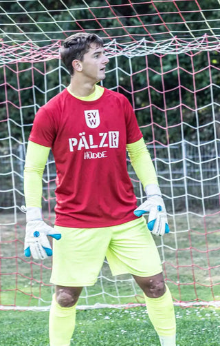 Ein männlicher Fußballtorwart trägt ein rotes Trikot, ein neon gelbes Unterhemd und Shorts sowie weiße und blaue Torwart-Handschuhe.