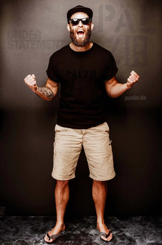 Ein Mann mit Bart, der ein schwarzes T-Shirt, beige Shorts und Flip-Flops trägt.