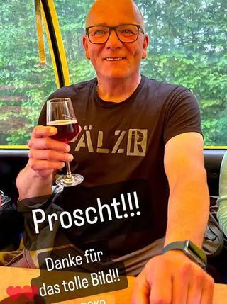 Ein Mann mit Brille und Glatze, der ein dunkles T-Shirt trägt und ein Glas Rotwein hält.