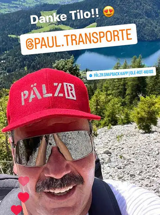 Ein lachender Mann trägt eine rote Mütze, reflektierende Sonnenbrillen und einen Oberlippenbart.