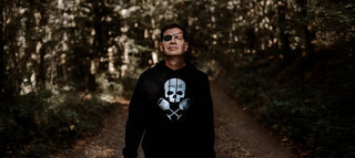 Mensch in einem schwarzen Hoodie mit einem Schädel- und gekreuzten Weinglas-Design, trägt eine Augenklappe.