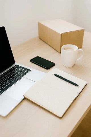 Offenes Laptop, ein leeres Notizbuch mit einem Stift, ein Smartphone, eine weiße Tasse und eine Pappkiste auf einem hellefarbenen Schreibtisch.