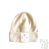 Cremeweiße gerippte Mütze mit einem Patch, der Trauben und das Wort ‚palzr‘ in Pink zeigt.