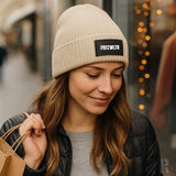 Beige Strickmütze mit ’frizwlr’-Patch, schwarze Reißverschlussjacke und grauer Pullover.