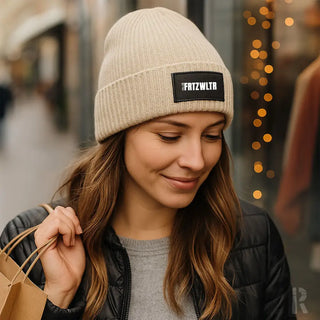 Beige Strickmütze mit ’frizwlr’-Patch, schwarze Reißverschlussjacke und grauer Pullover.