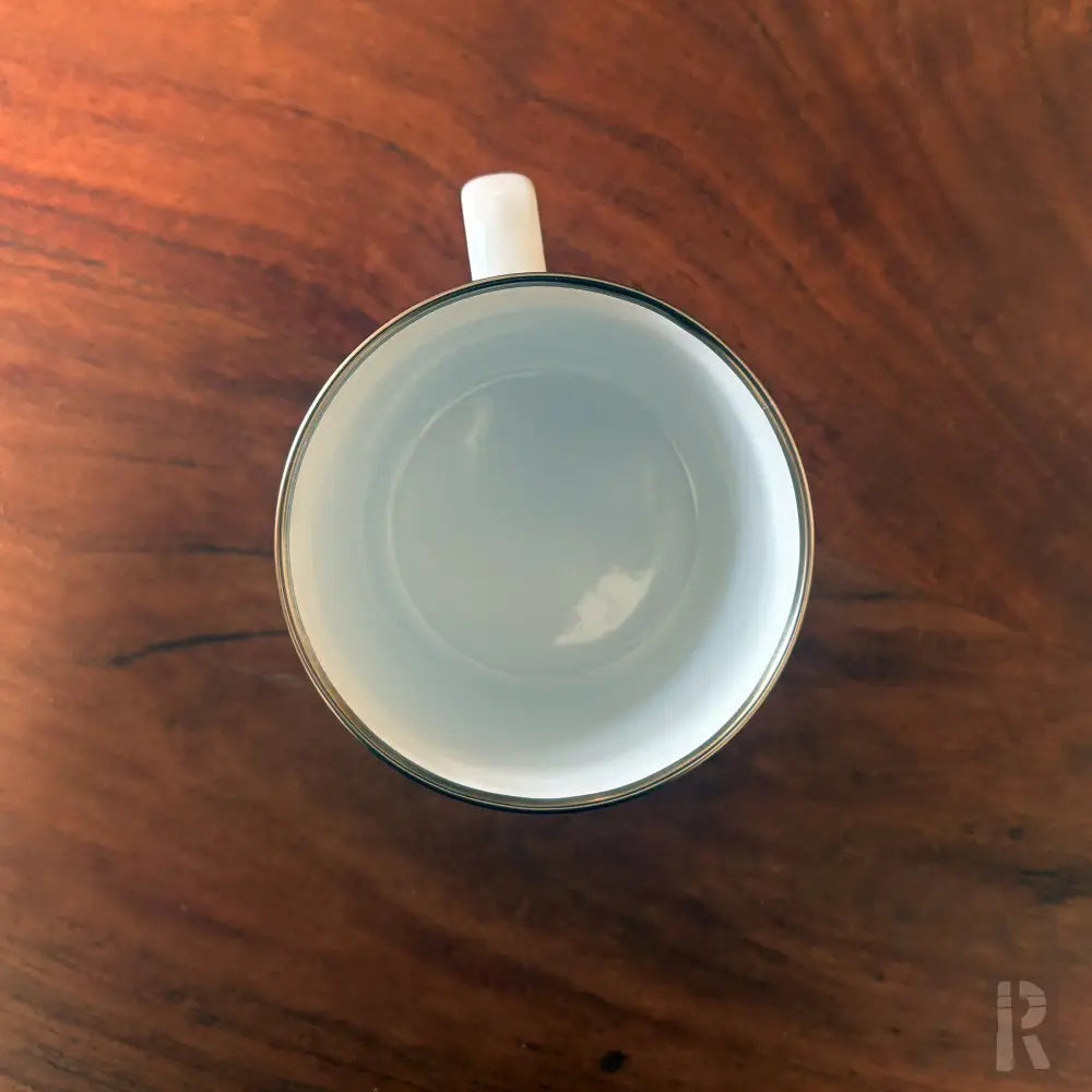 Weißer Emaille-Kaffeebecher mit schwarzem Rand und Griff, rustikaler Holztisch-Hintergrund.