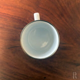 Weißer Emaille-Kaffeebecher mit schwarzem Rand und Griff, rustikaler Holztisch-Hintergrund.