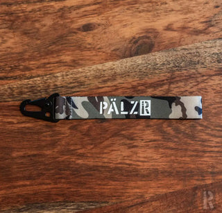 Camouflage-Schlüsselanhänger mit ’pälzr’-Marke in Weiß auf schwarzem Hintergrund.