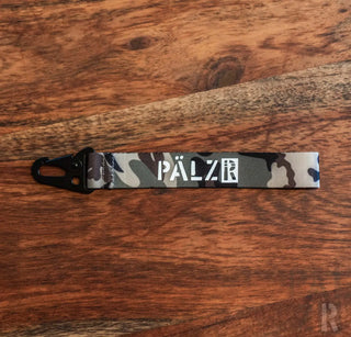 Camouflage-Schlüsselanhänger mit ’pälzr’-Marke in Weiß auf schwarzem Hintergrund.