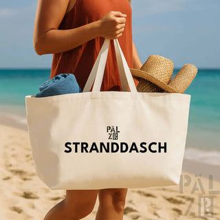 Weiße Tragetasche mit schwarzem Text ’stranddasch’ und Strohhut darin, Strandessentials.