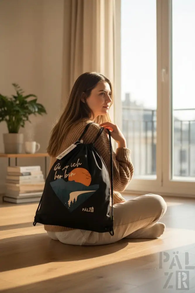 Schwarzer Rucksack mit Kordelzug und einem Grafikdesign, das eine orange Sonne über Bergen zeigt und den deutschen Satz ’die wo ich her bin’ in weißer Schrift gedruckt hat, zusammen mit dem Markennamen ’palz’ am unteren Rand.
