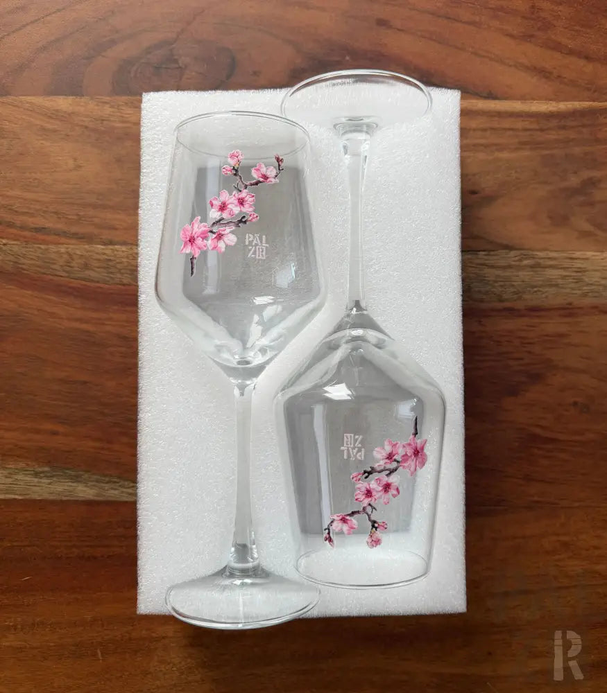 Ein Paar klare Glas-Weingläser mit zarten rosa Kirschblüten-Designs und ’pal zir’-Marke, eingebettet in weißer Schaumverpackung.