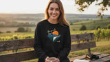 Schwarzer Sweatshirt mit langen Ärmeln mit einem grafischen Aufdruck einer orangefarbenen Sonne über einer weißen Bergsilhouette und dem Text „da wo ich her bin“ darüber, mit einem kleinen Logo in der unteren rechten Ecke.
