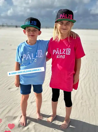 Zwei Kinder tragen passende „PÄLZER Kind’ T-Shirts und Hüte an einem Strand.