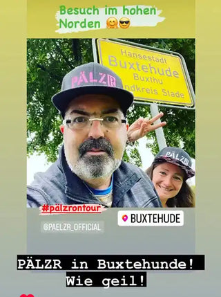 Zwei Personen tragen passende „PÄLZR„-Mützen und Brillen und posieren vor einem gelben Ortsschild für „Hansestadt Buxtehude„.